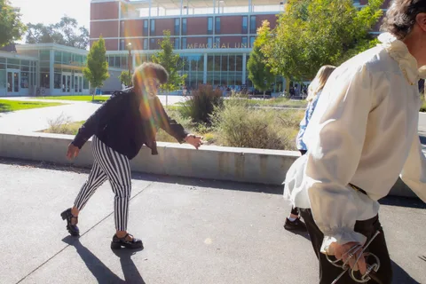 A woman in striped trousers bends double in a zombie stagger as a man in a ruffled shirt and sword walks past, sun flaring across the frame.