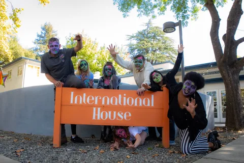 The group drapes itself over the orange International House sign, one person kneeling underneath and reaching through the gap.