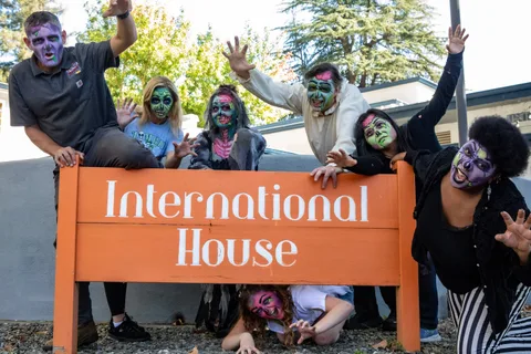 Zombies crowd both ends of the orange International House sign with hands raised, one crouched low behind the boards.