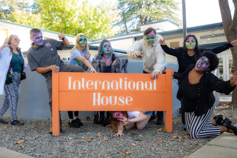 The whole group lines up along the orange International House sign in front of the building's grey wall.