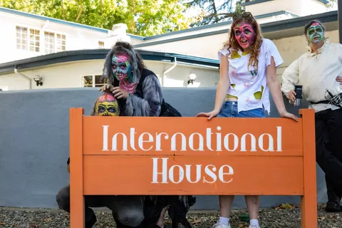 Two zombies lean on the International House sign beside a woman in a stained t-shirt, with another face peering over the boards.