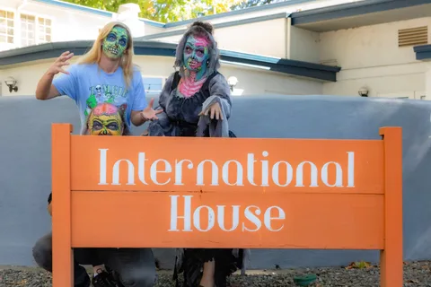 A woman in a Zombie Bike Parade t-shirt and a zombie in a grey shroud pose behind the International House sign, a third face peeking over the top.