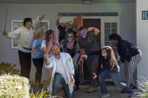 Zombies spill out of the International House Davis entrance, a man in a lab coat kneeling in front with one arm reaching toward the camera.