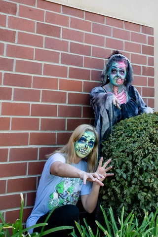 A woman in a Zombie Bike Parade t-shirt crouches at a brick wall reaching toward the camera, with a zombie in a grey shroud rising behind the hedge.