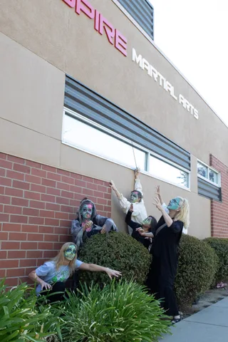 Zombies crouch behind the hedges while one reaches up with a thin sword toward the Inspire Martial Arts sign.