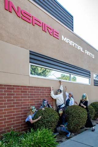A man in a ruffled shirt stretches a sword up toward the Inspire Martial Arts letters while five zombies lurk in the hedge below.